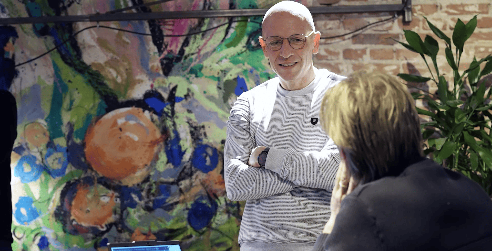 Man in glasses and light sweater smiling while conversing with woman in front of colorful abstract artwork